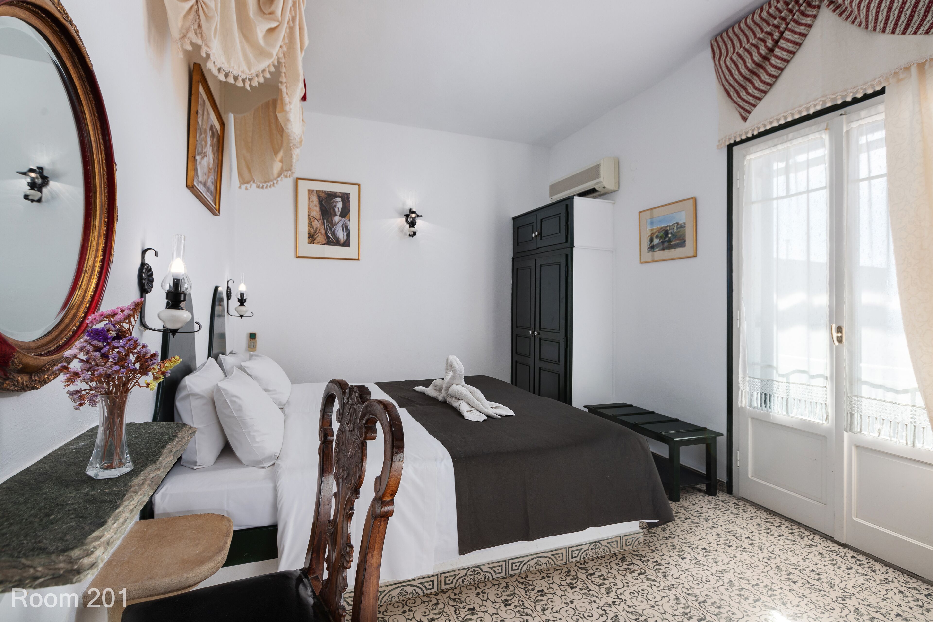 A bedroom with a large bed, a wooden chair, a desk with a vase of flowers, a mirror, and a wardrobe.