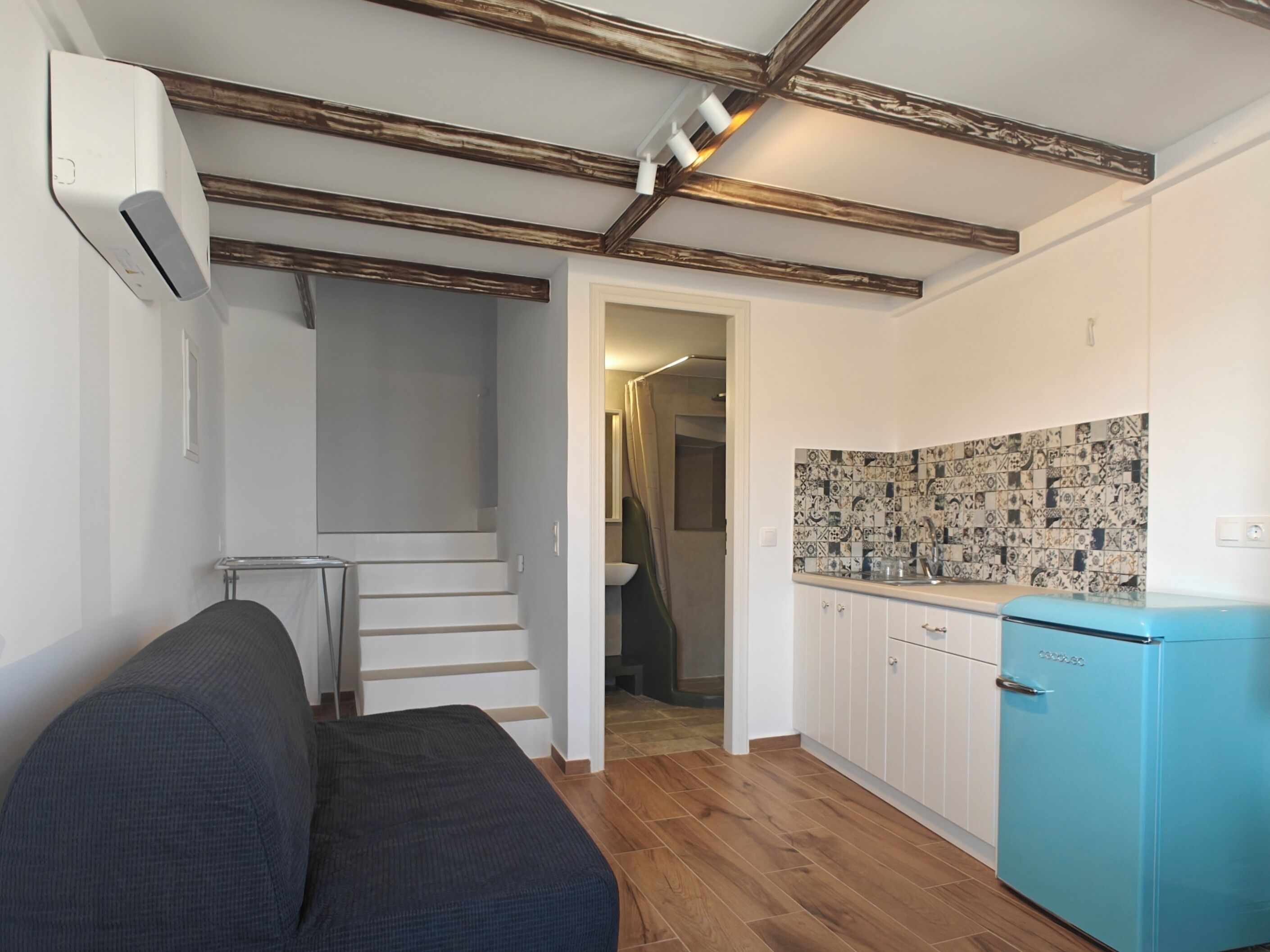 A compact living space with a sofa, a kitchenette with patterned tiles, a blue refrigerator, and a staircase leading to another room.
