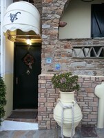 Un edificio de piedra con una puerta oscura, un toldo blanco con un logotipo, una planta en maceta y un cartel pequeño.