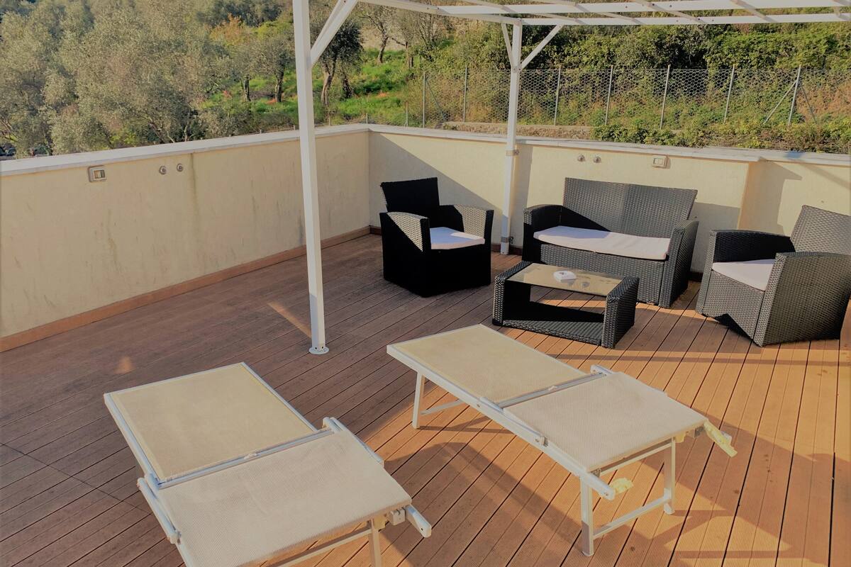 Un espace extérieur aménagé, partiellement abrité, avec des fauteuils et une table, entouré d’une terrasse en bois et d’une pergola blanche.