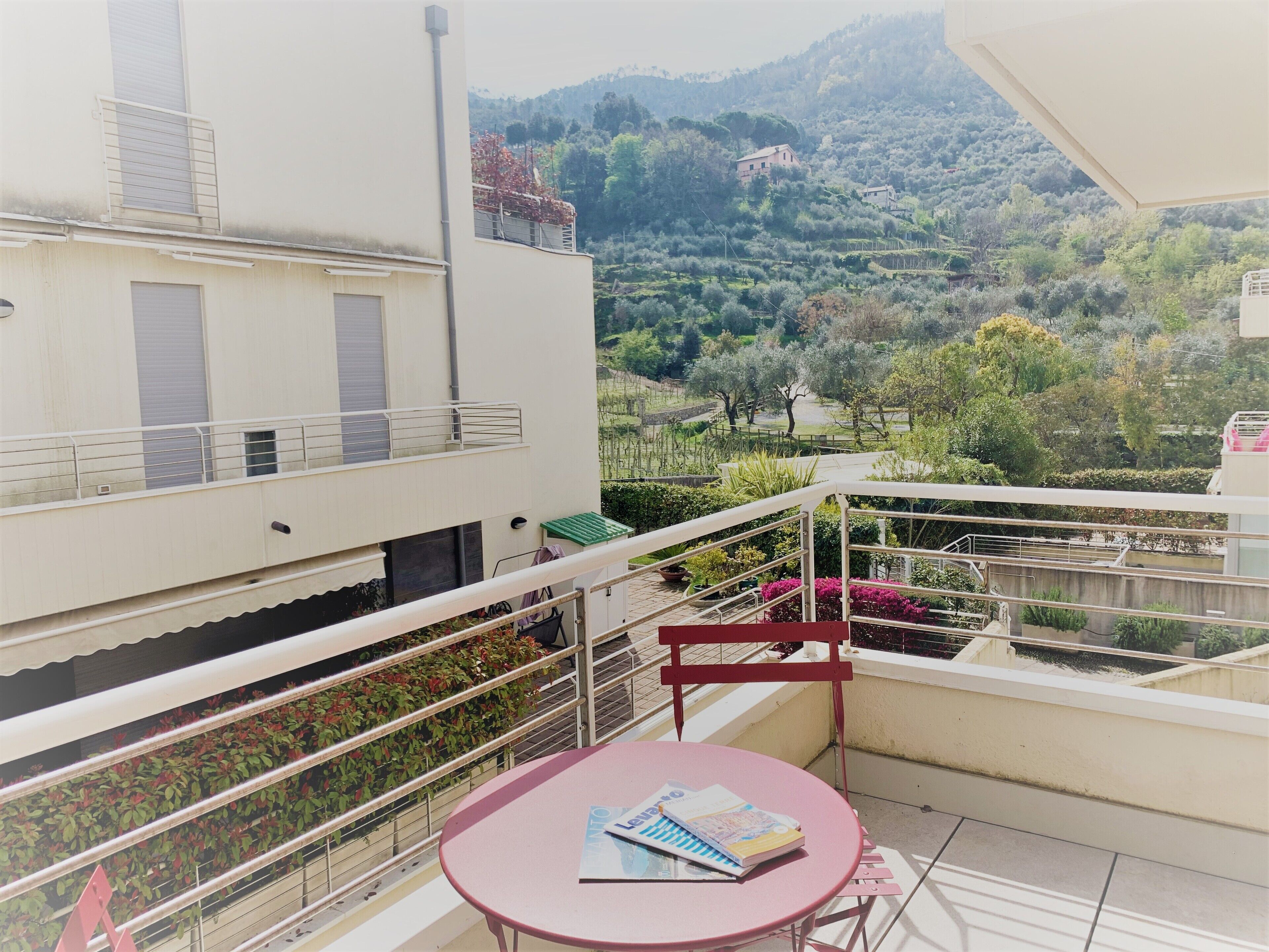 Un balcon avec une table et une chaise, donnant sur un paysage montagneux parsemé d’arbres et sur lequel on aperçoit un bâtiment au loin.