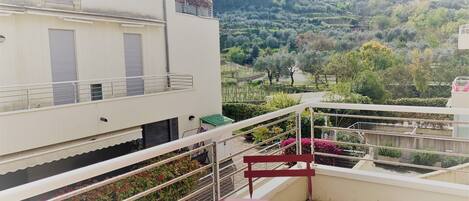 A balcony with a table and chair, overlooking a mountainous landscape with trees and a building in the distance.