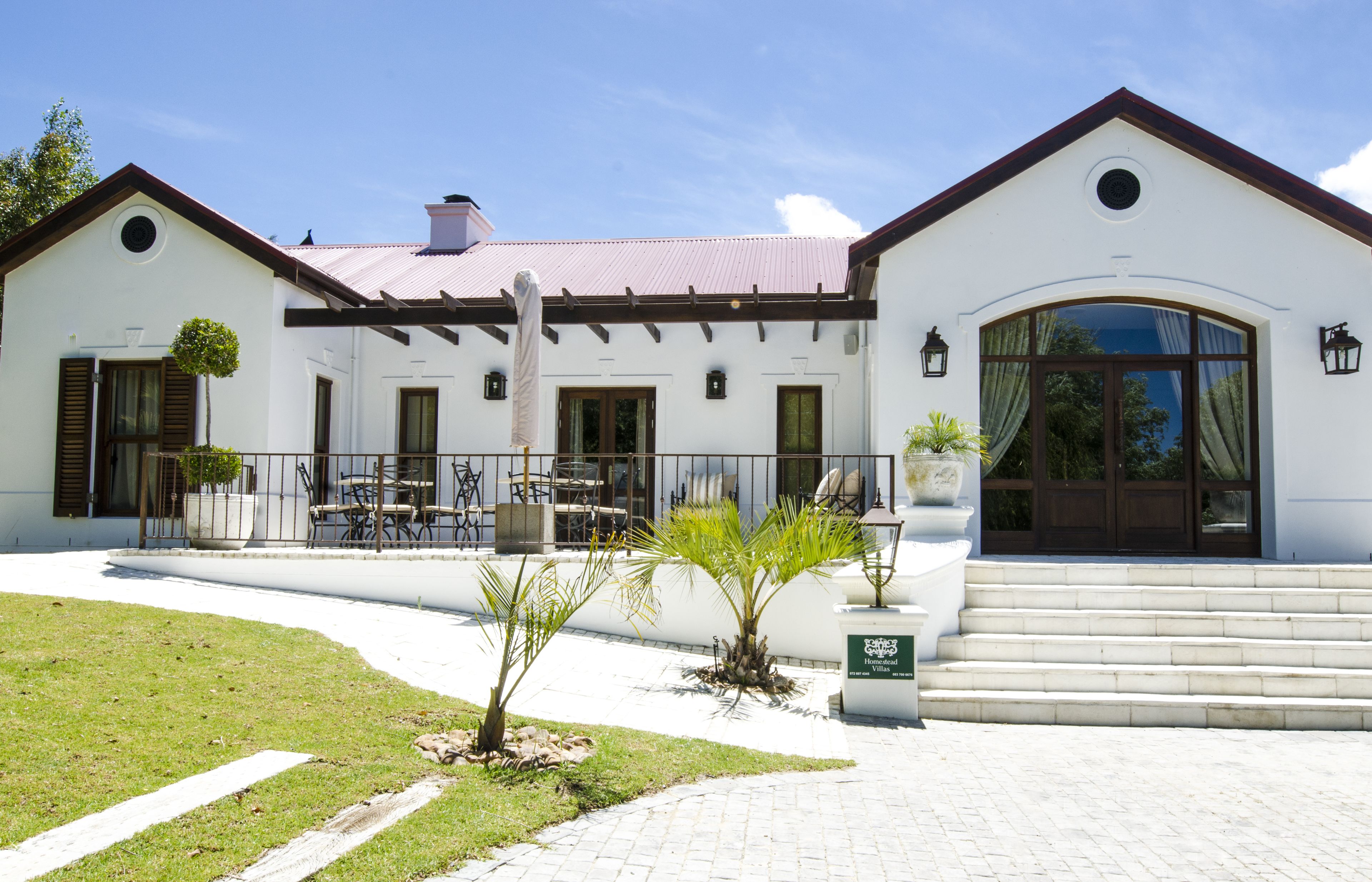 A two-story building with a white facade, a red roof, and a large arched entrance. There are outdoor seating areas with tables and chairs, and a small palm tree in the foreground.