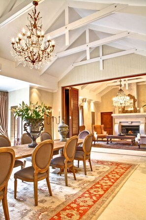 A spacious dining area with a high ceiling, chandeliers, and a fireplace. - Homestead Villas (Cape Town)