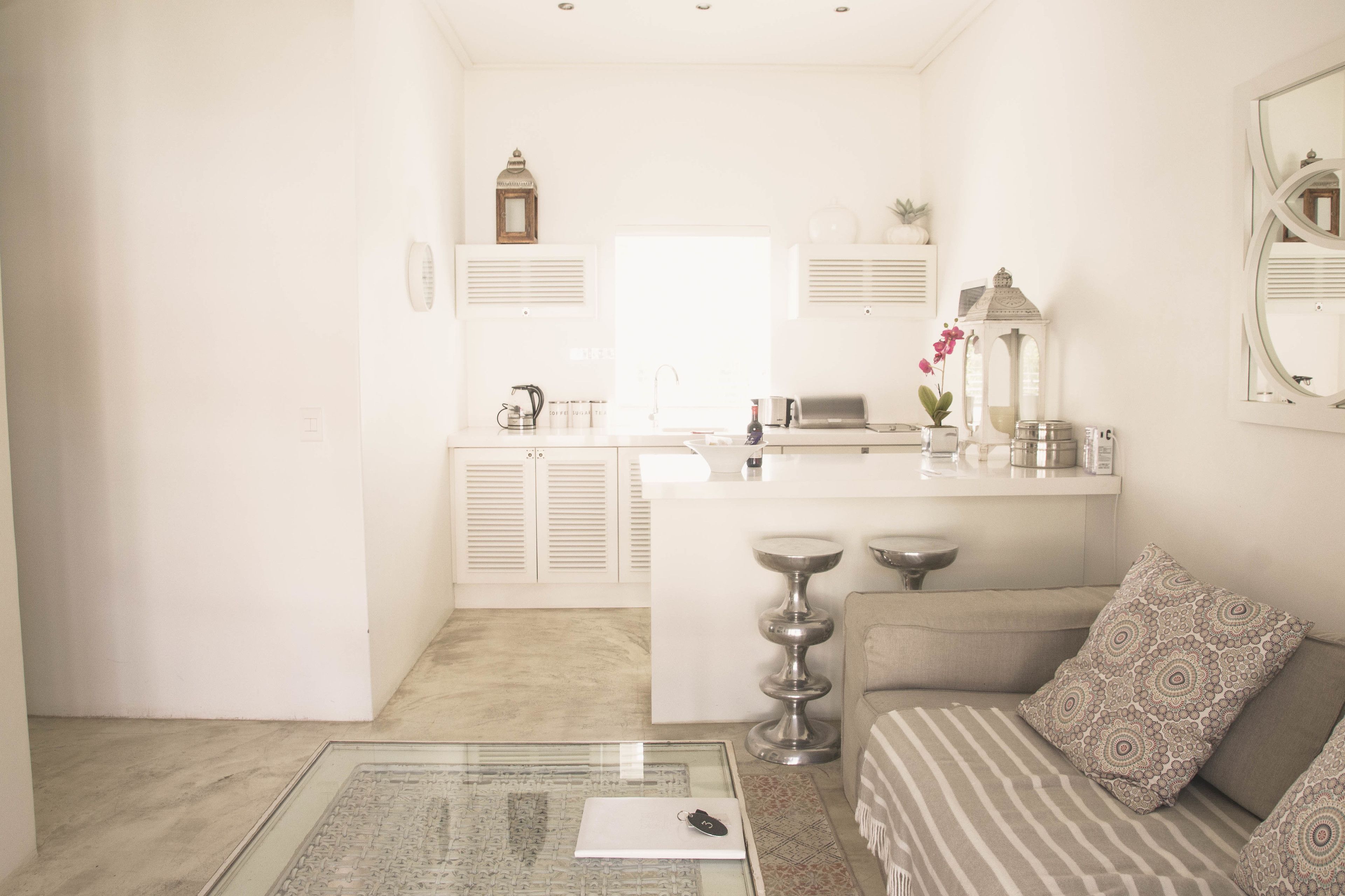 A bright, minimalist living room with a glass coffee table, a sofa, and a kitchen area in the background.