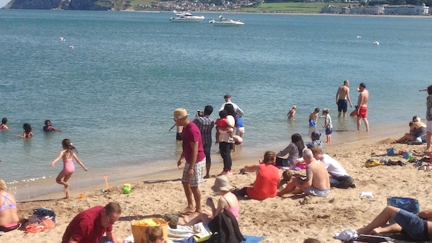Una playa con gente tomando el sol y jugando en la arena, barcos en el agua y colinas al fondo.