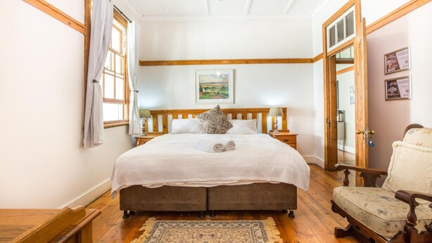 A bedroom with a large bed, wooden headboard, a nightstand, a chair, and a view of a landscape through a window. - Coweys Corner (Durban)