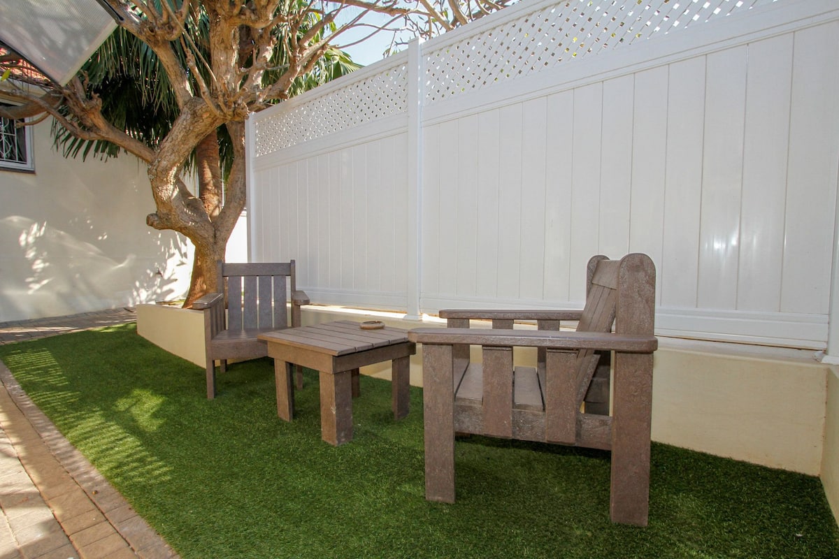 An outdoor seating area with a wooden table and chairs, a tree, and a white fence.