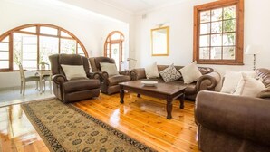 A spacious living room with a wooden floor, brown leather sofas, a coffee table, and large windows with wooden frames. - Coweys Corner (Durban)