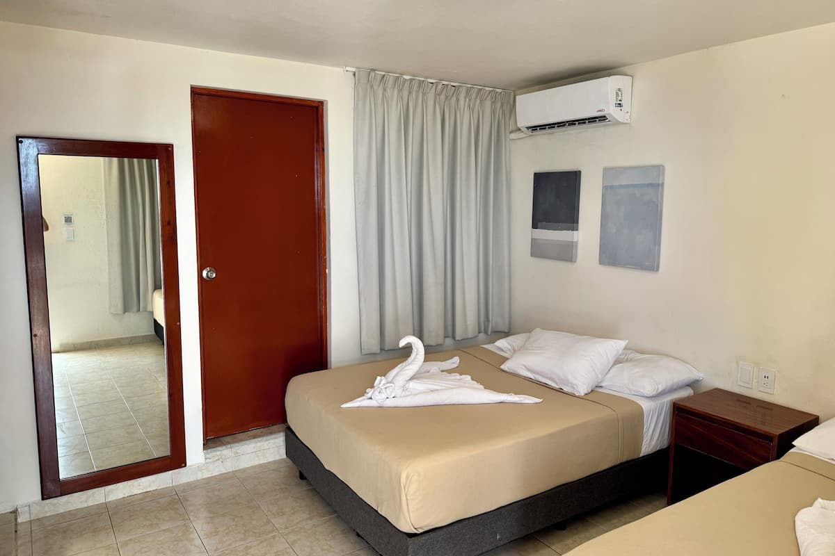 A hotel room with a bed, a wooden nightstand, a wall-mounted air conditioner, and a door leading to another room.