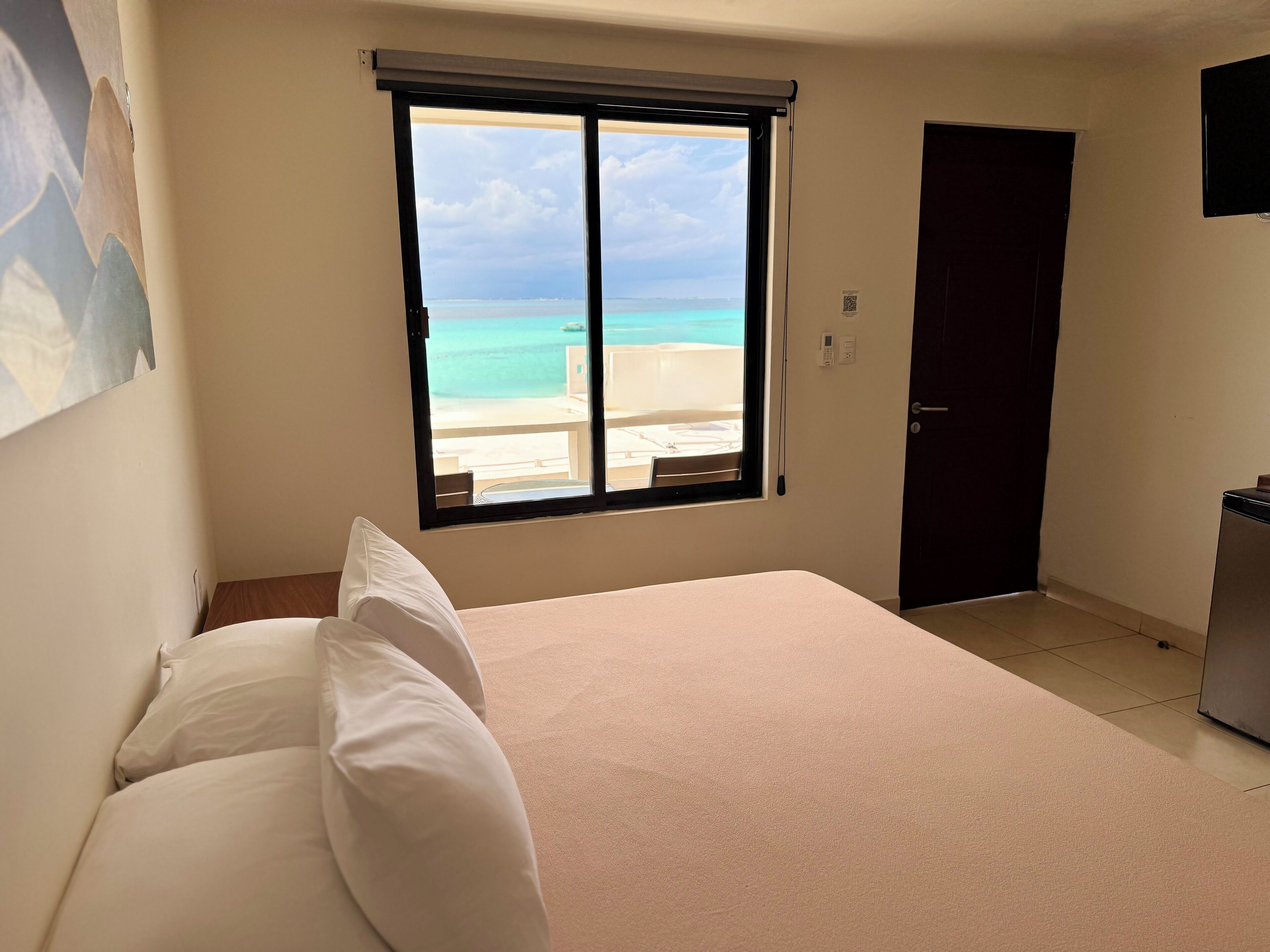 A hotel room with a bed, a window with a view of the beach, a door, and a small table.
