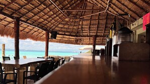 Un área de comedor al aire libre con techo de paja, mesas y sillas de madera, con vista a una playa de aguas turquesas cristalinas.