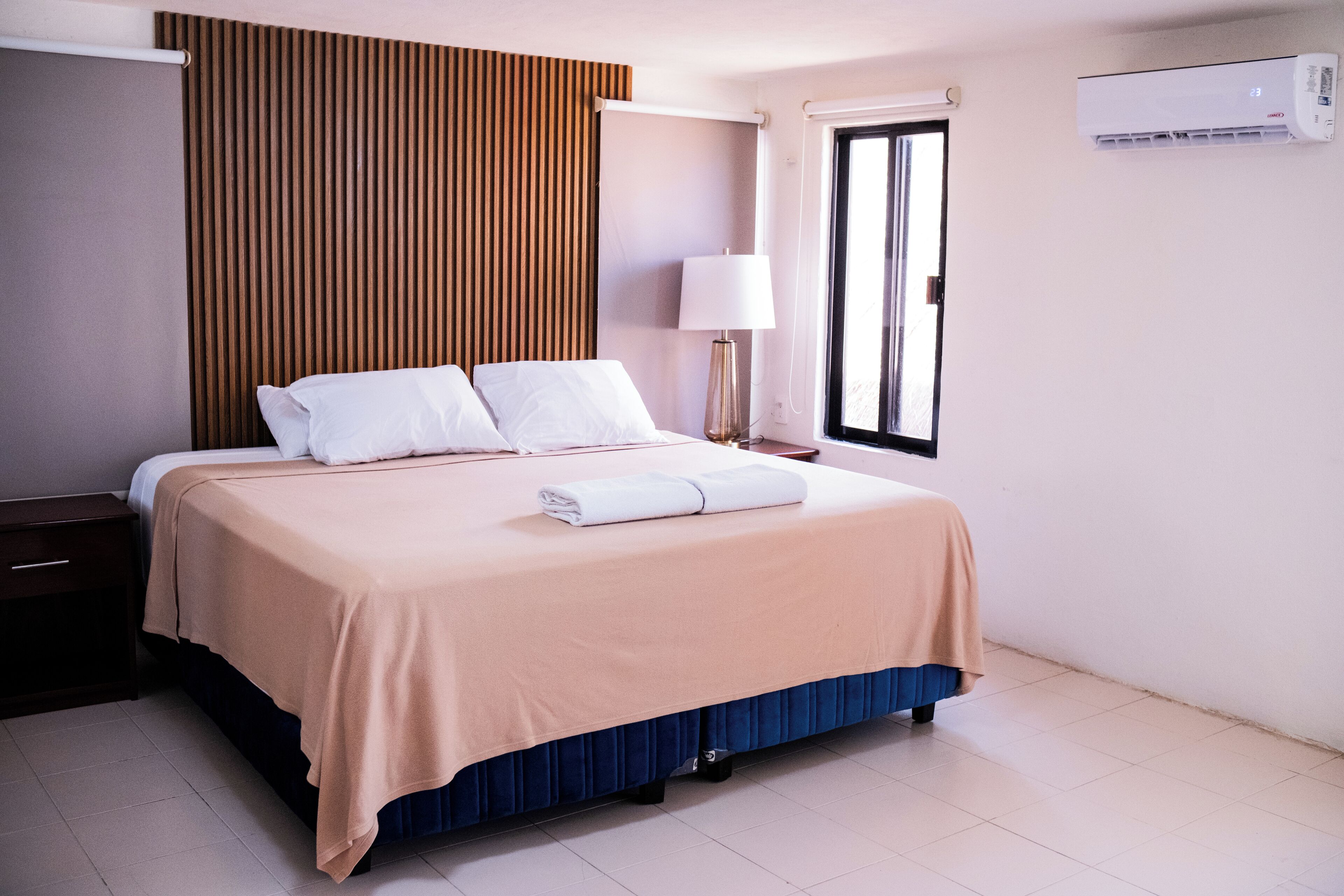 A neatly made bed with a beige bedspread and a blue skirted base, a bedside table with a lamp, and an air conditioning unit on the wall.