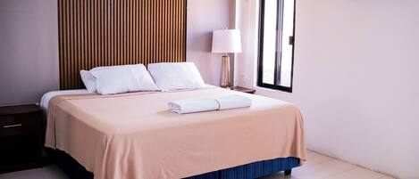 A neatly made bed with a beige bedspread and a blue skirted base, a bedside table with a lamp, and an air conditioning unit on the wall.