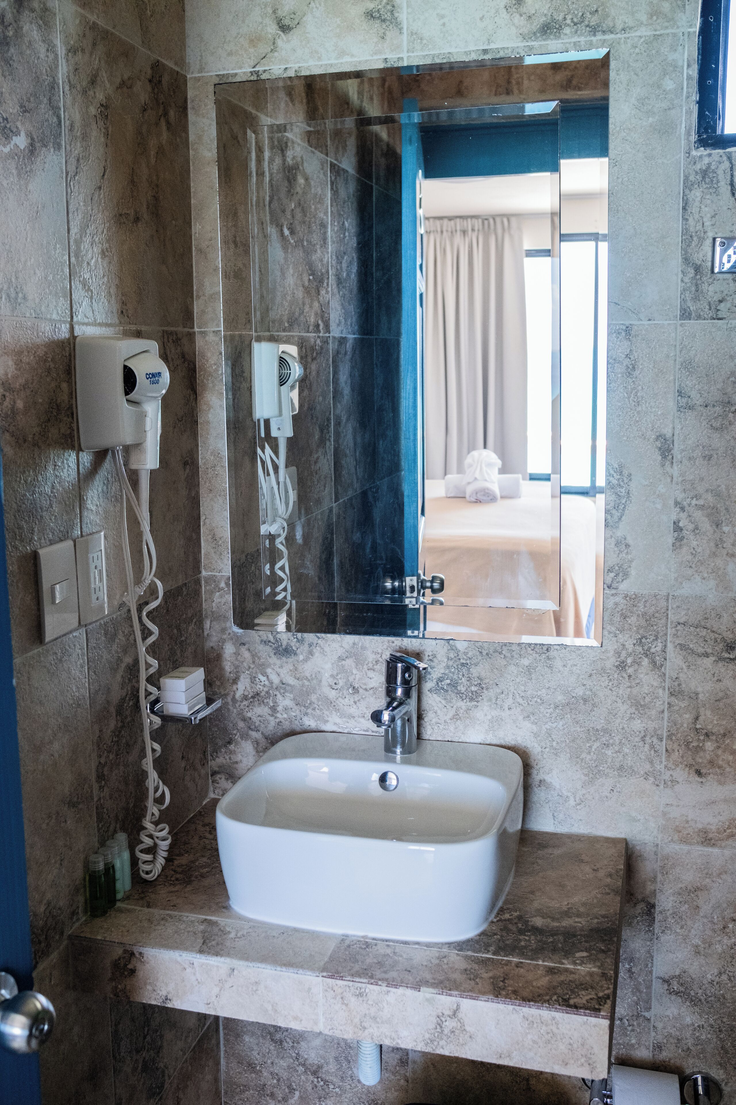 A bathroom with a marble sink, a hairdryer, and a mirror reflecting a bed with a towel.
