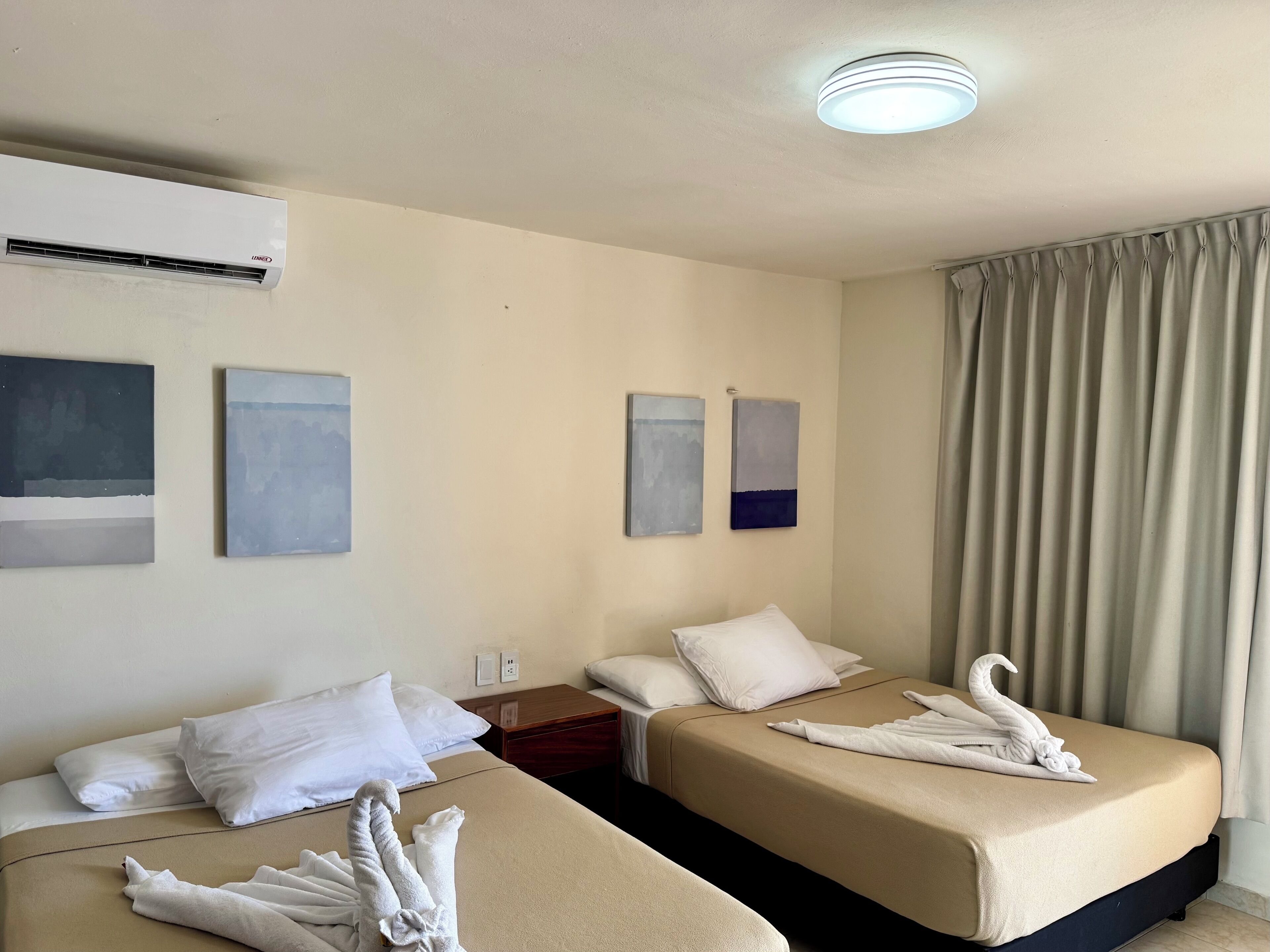 Una habitación de hotel con dos camas, ropa de cama color beige, toallas blancas y obras de arte en las paredes.