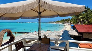 Un patio junto a la playa con una mesa y sillas, un paraguas grande y vistas al océano y a gente disfrutando en la playa.