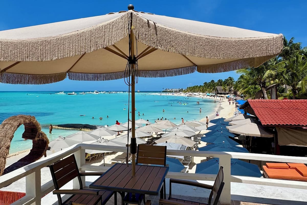 A beachside patio with a table and chairs, a large umbrella, and a view of the ocean and beachgoers.