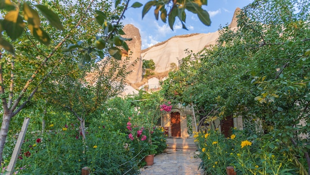 A stone pathway leading to a rustic building with a wooden door, surrounded by lush greenery and flowering plants. - Roc Of Cappadocia (Nevsehir)