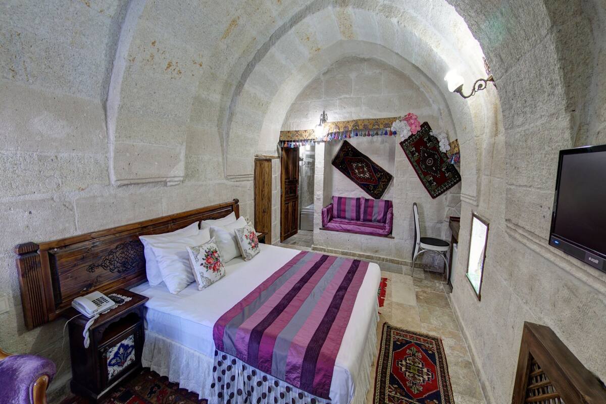 A room with a bed, a wooden headboard, a nightstand, a TV, and a stone wall with an arched ceiling.