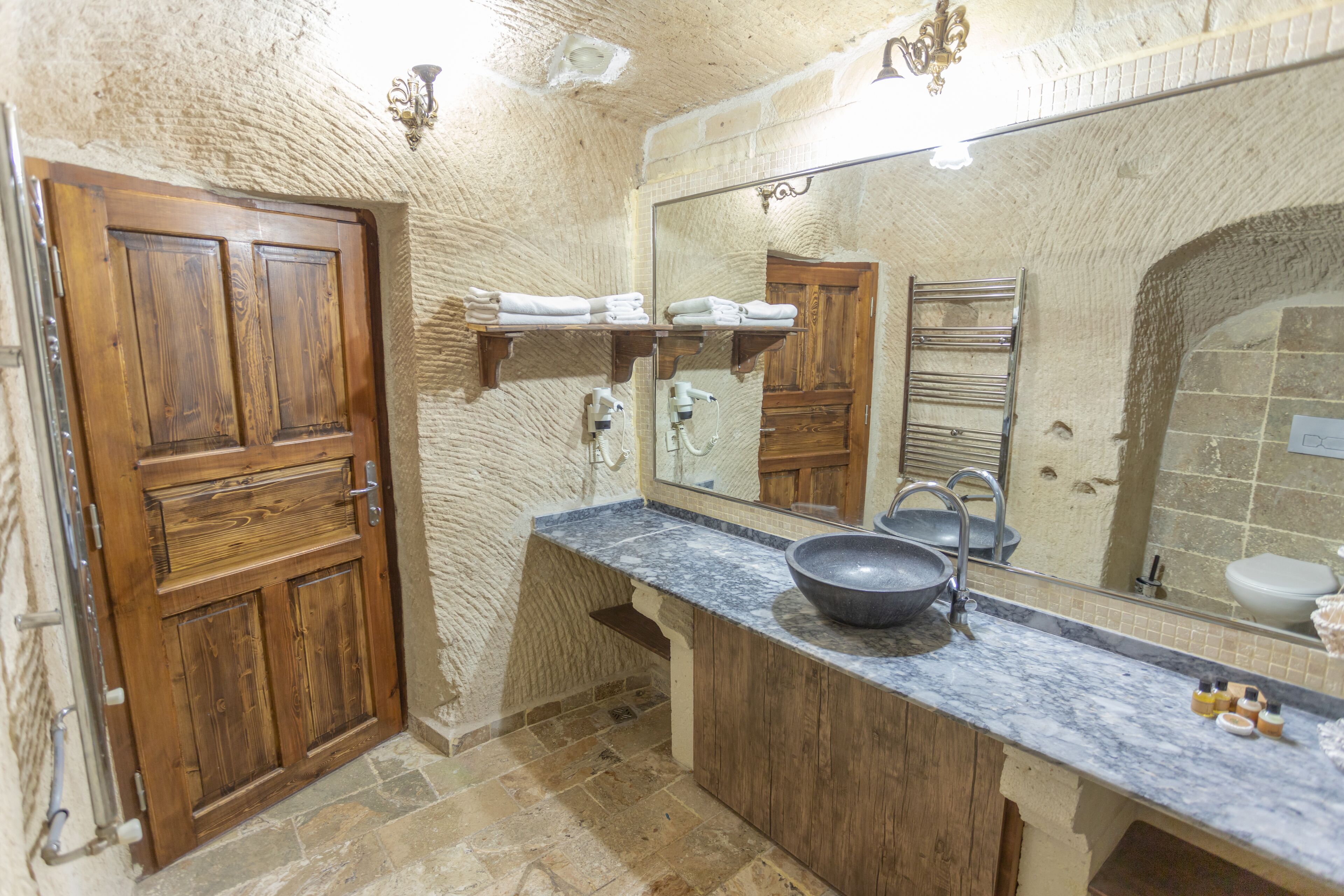 Un baño con puerta de madera, un espejo grande, encimera de mármol y suelo de piedra.