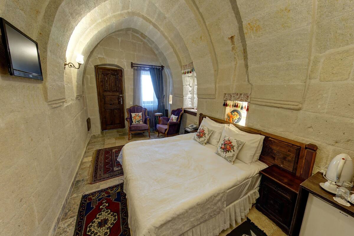 A bedroom with a large bed, a television mounted on the wall, and a stone archway leading to another room.