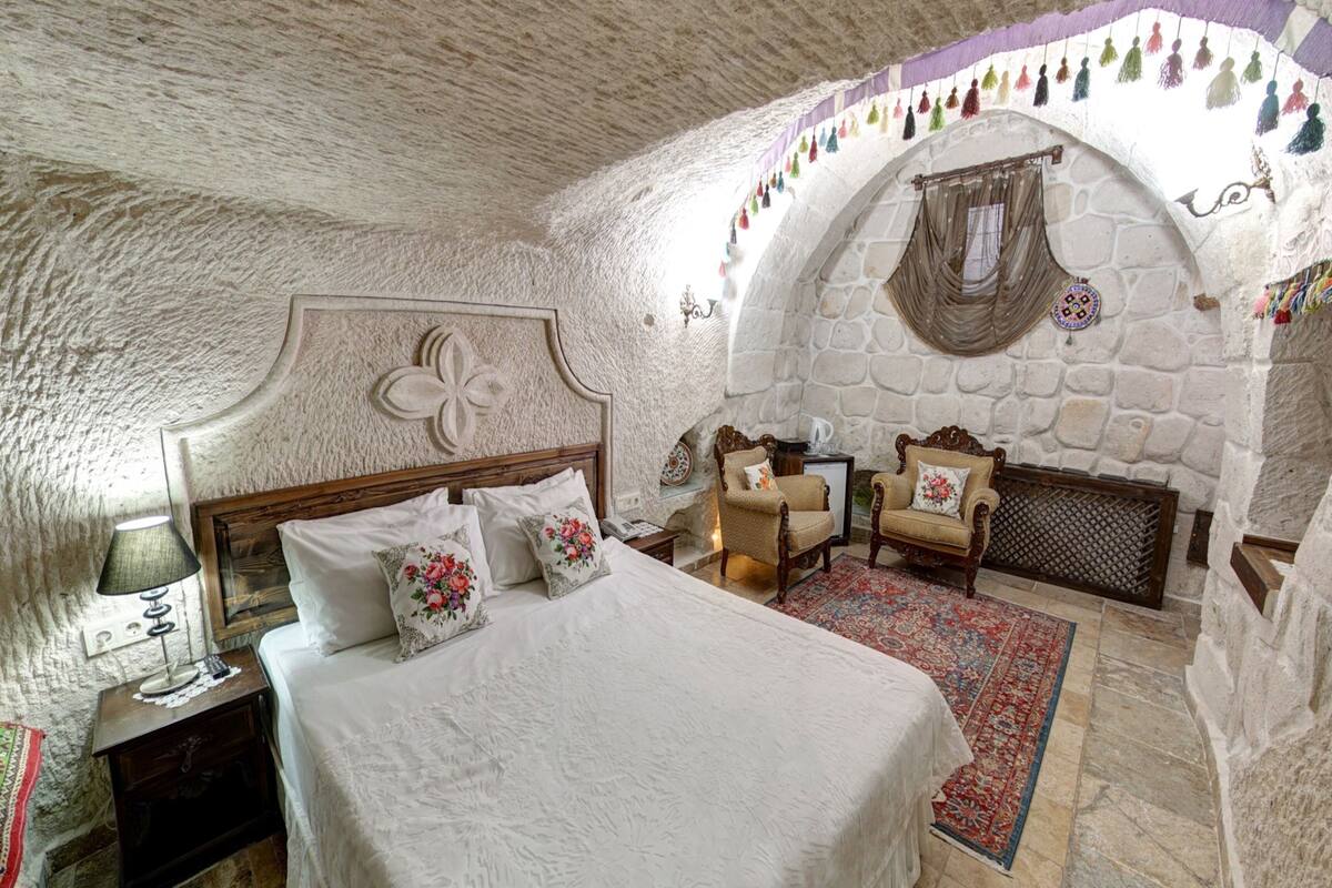 A bedroom with a stone wall, a bed with white linens, a wooden headboard, a chair, and a small table with a lamp.