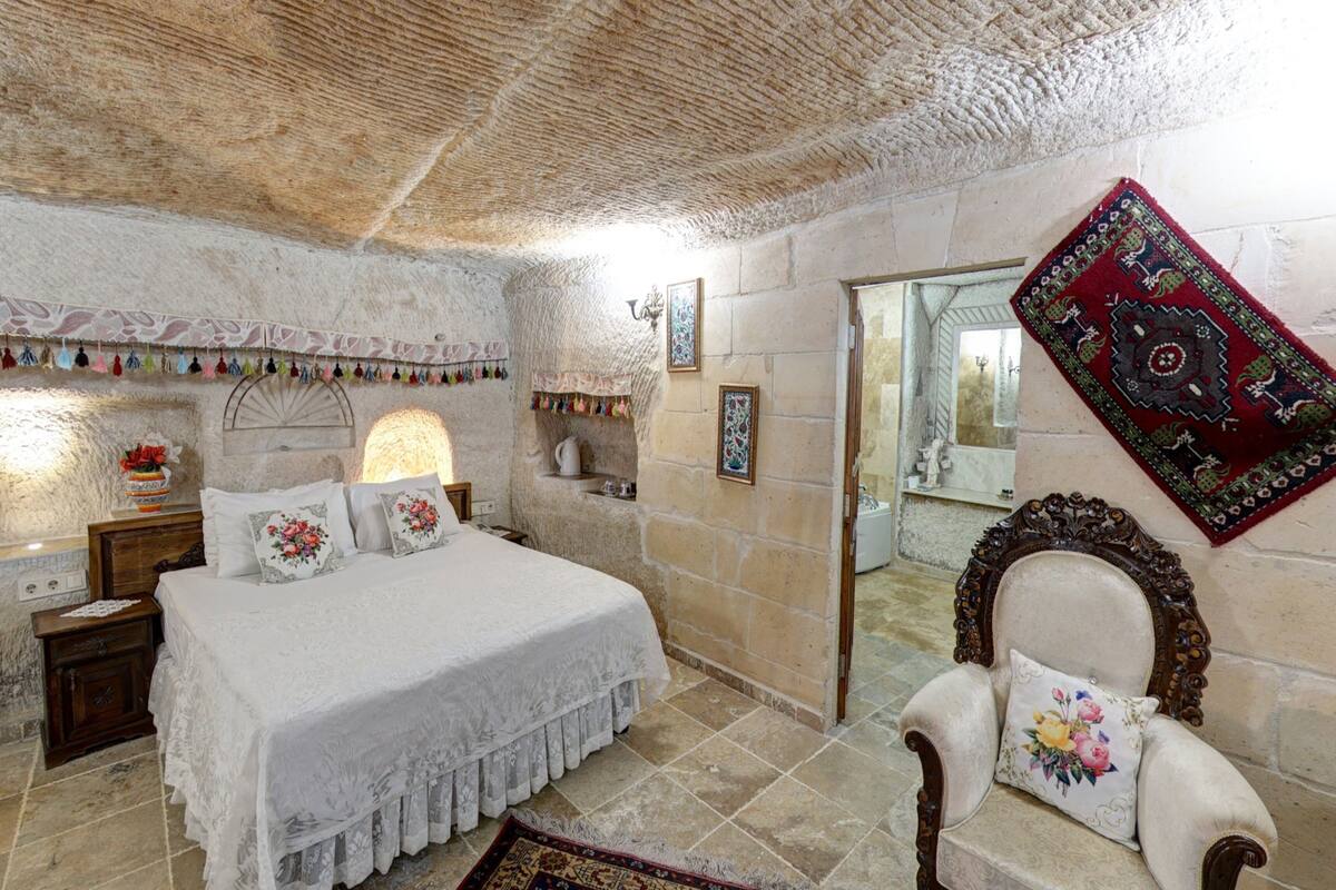 A bedroom with a stone wall, a bed with white bedding, a wooden nightstand, a chair with a floral pillow, and a red patterned rug.