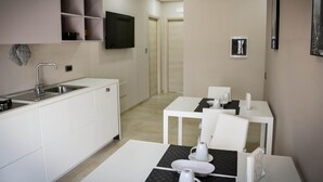 A modern kitchen with white cabinets, a built-in sink, and a flat-screen TV mounted above the counter. There are two white dining tables with black placemats and white chairs. - Brain Rooms & Suite (Pagani)