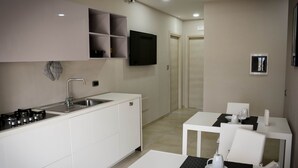 A modern kitchen with white cabinets, a built-in stove, and a flat-screen TV. There is a dining area with a table and chairs. - Brain Rooms & Suite (Pagani)