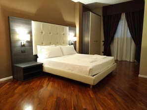 A modern bedroom with a large bed, a nightstand, a wall-mounted lamp, and a view of the outside through a window with curtains. - Brain Rooms & Suite (Pagani)