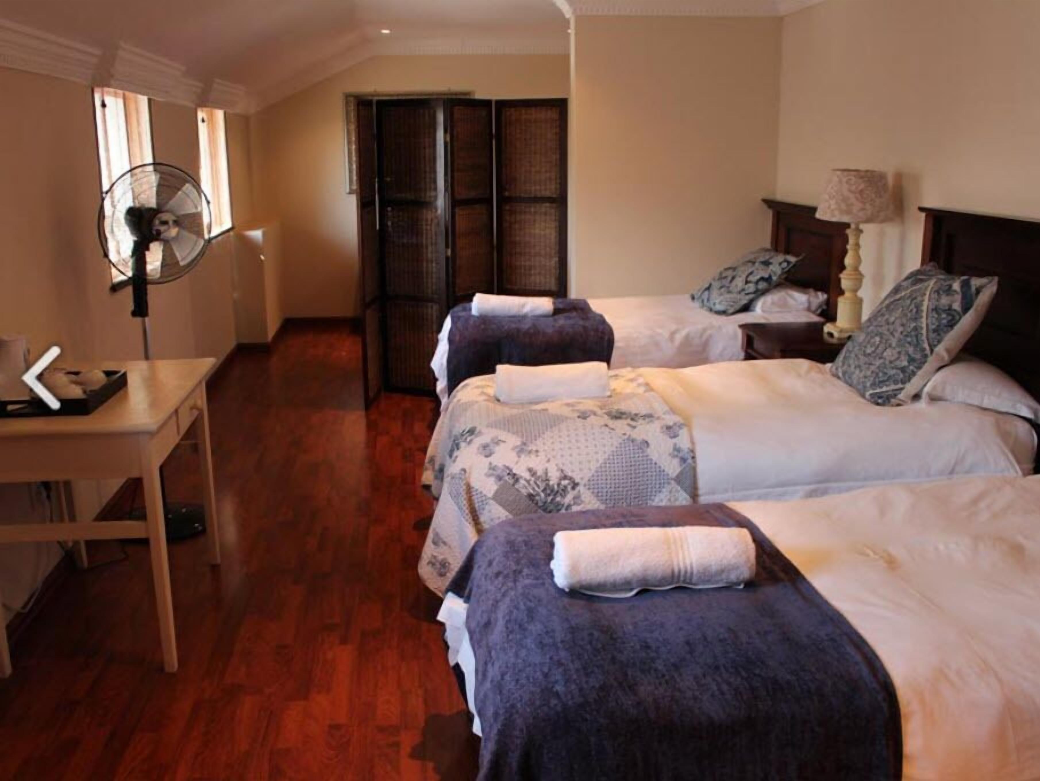 Quarto de hotel com duas camas, uma escrivaninha, uma cadeira, um ventilador e uma janela.