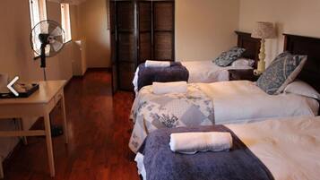 Quarto de hotel com duas camas, uma escrivaninha, uma cadeira, um ventilador e uma janela.