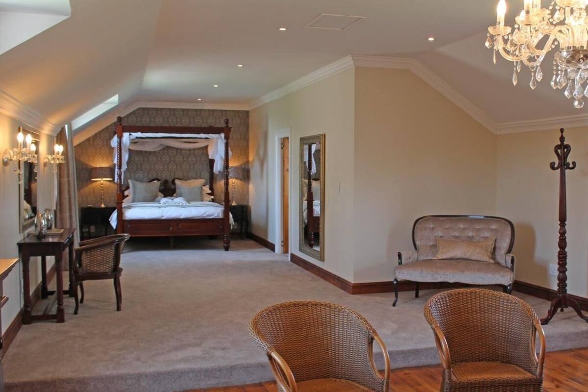 A spacious bedroom with a large bed, a sitting area with wicker chairs, a chandelier, and a view of the outdoors.