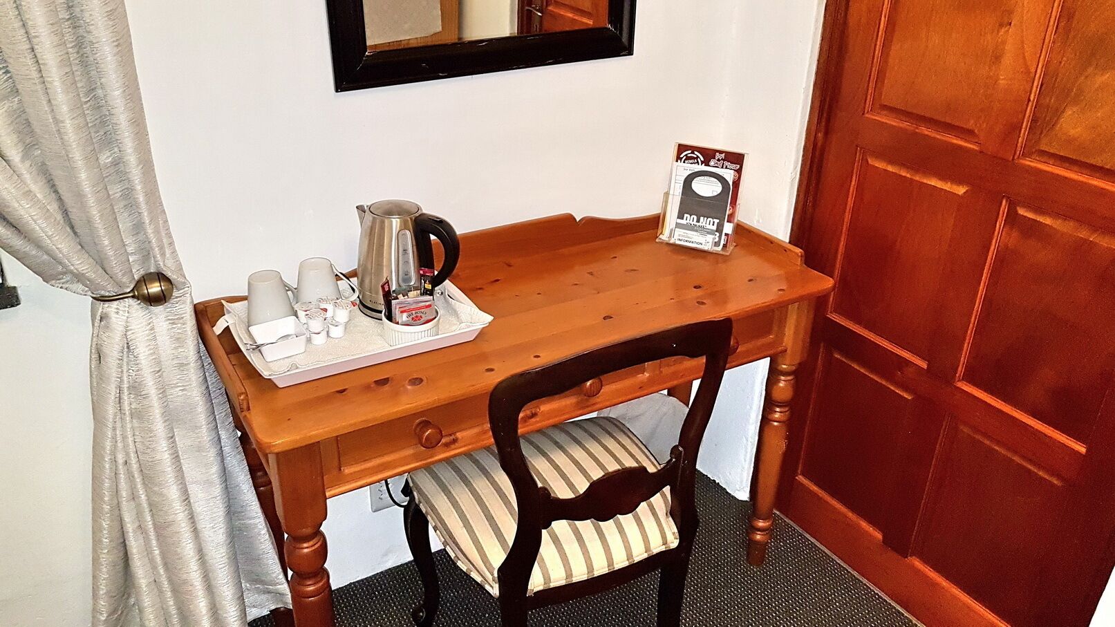 Un bureau en bois, avec une bouilloire, des tasses et un plateau contenant des articles de toilette. Une chaise avec un coussin à rayures est placée devant le bureau.