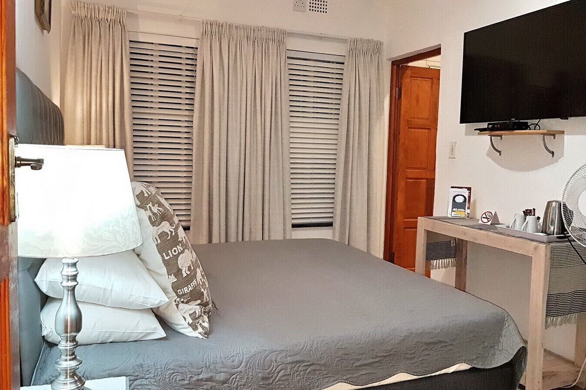 Quarto de hotel com cama, escrivaninha, televisão, ventoinha e porta.