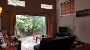 A living room with a sofa, a coffee table, a TV, and a fan. There are windows with blinds and a view of greenery outside. - Six Valk Avenue Guest House (Sandton)