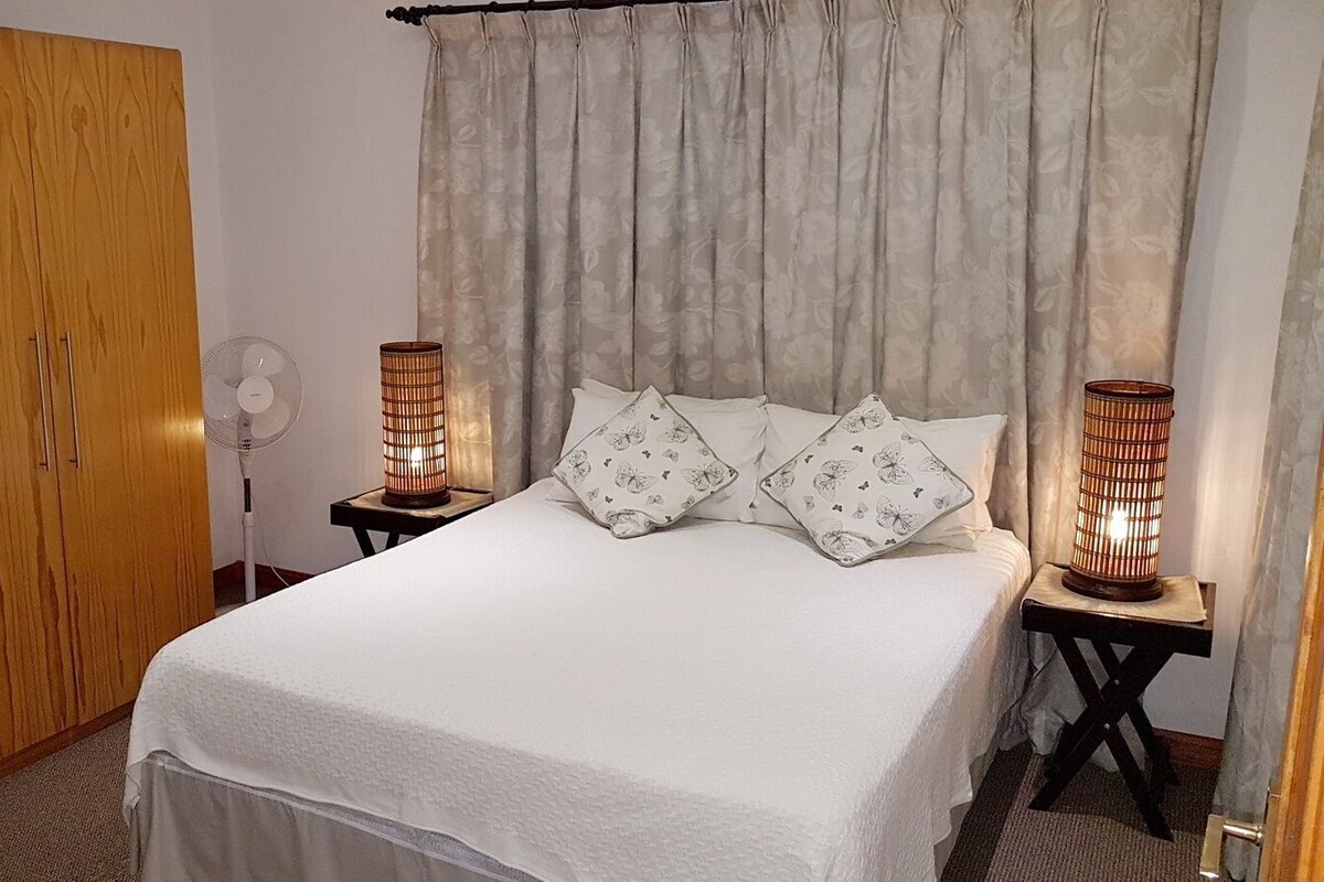 Quarto com uma cama, duas mesas de cabeceira com abajures, um guarda-roupa de madeira e um ventilador.