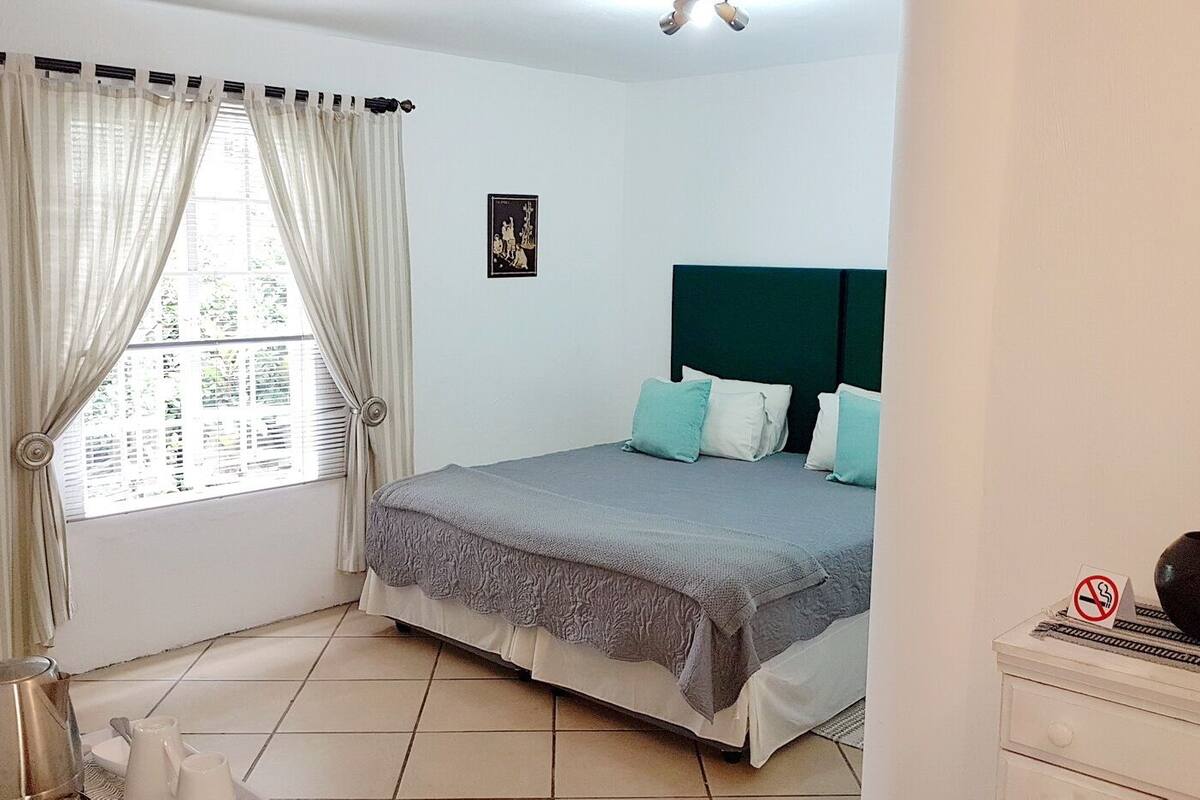 Une chambre à coucher avec un lit, des rideaux, une fenêtre, un mur orné de tableaux et un vase posé sur une commode.