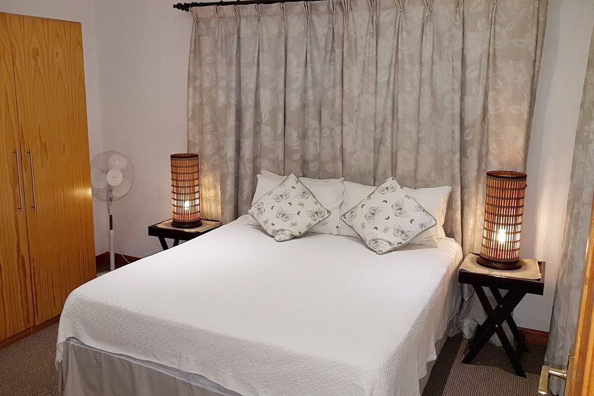 Une chambre à coucher comprenant un lit, deux tables de chevet avec des lampes, une armoire en bois et un ventilateur sur pied.