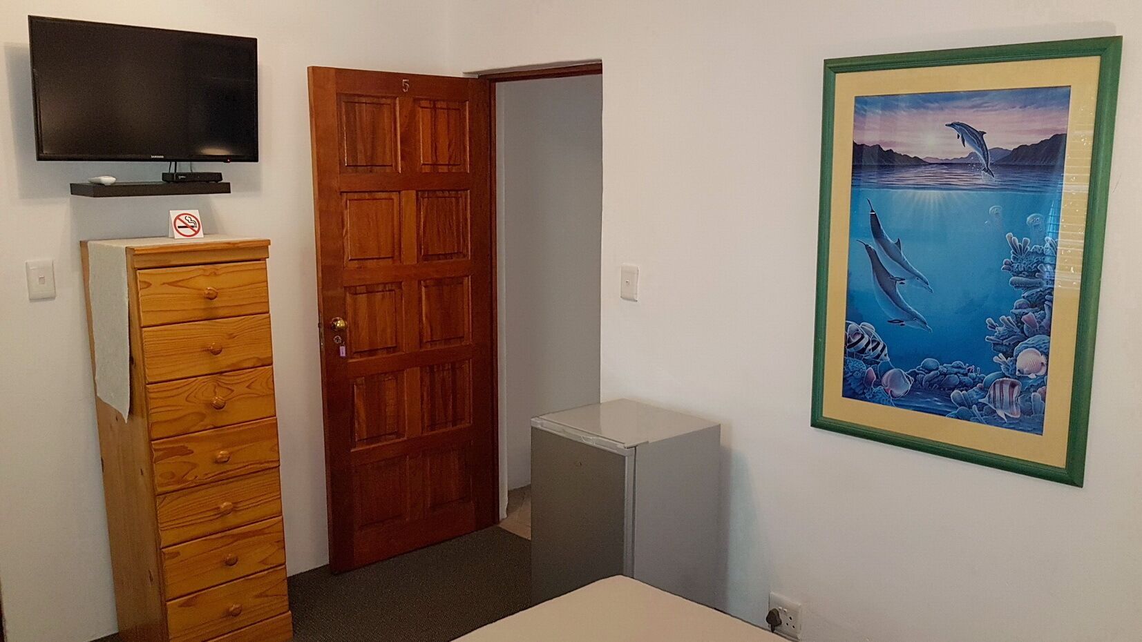 A room with a wooden dresser, a closed door, a small gray cabinet, a mounted television, and a framed picture of dolphins.