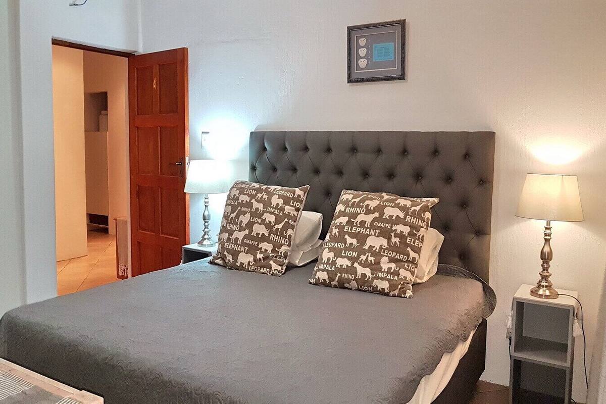 Un lit bien fait, avec une couette grise et deux coussins à motifs, une lampe de chevet et un tableau encadré accroché au mur.