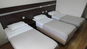 Une chambre d’hôtel avec trois lits simples, chacun doté d’un matelas et d’un oreiller blancs, d’une tête de lit en bois et d’une table de chevet avec une lampe.