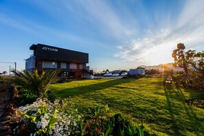 A modern building with a sign reading "JOYUAM" in a well-maintained grassy area with flowers and a clear sky. - Joyuam Pension (Seogwipo)