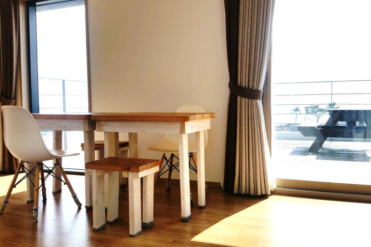 A room with a wooden table and a white chair, a window with curtains, and a view of a balcony with a table and chairs.