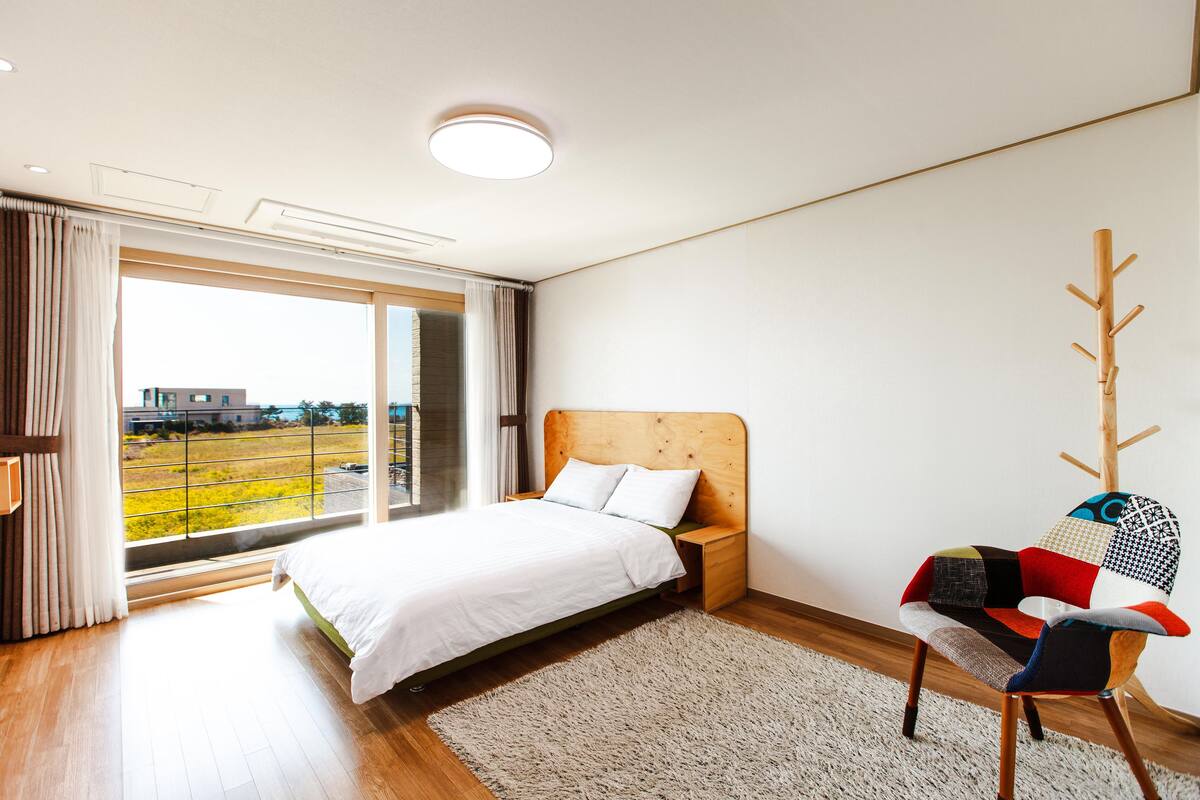 A modern bedroom with a large bed, a wooden nightstand, a colorful chair, and a view of a green landscape through a large window.