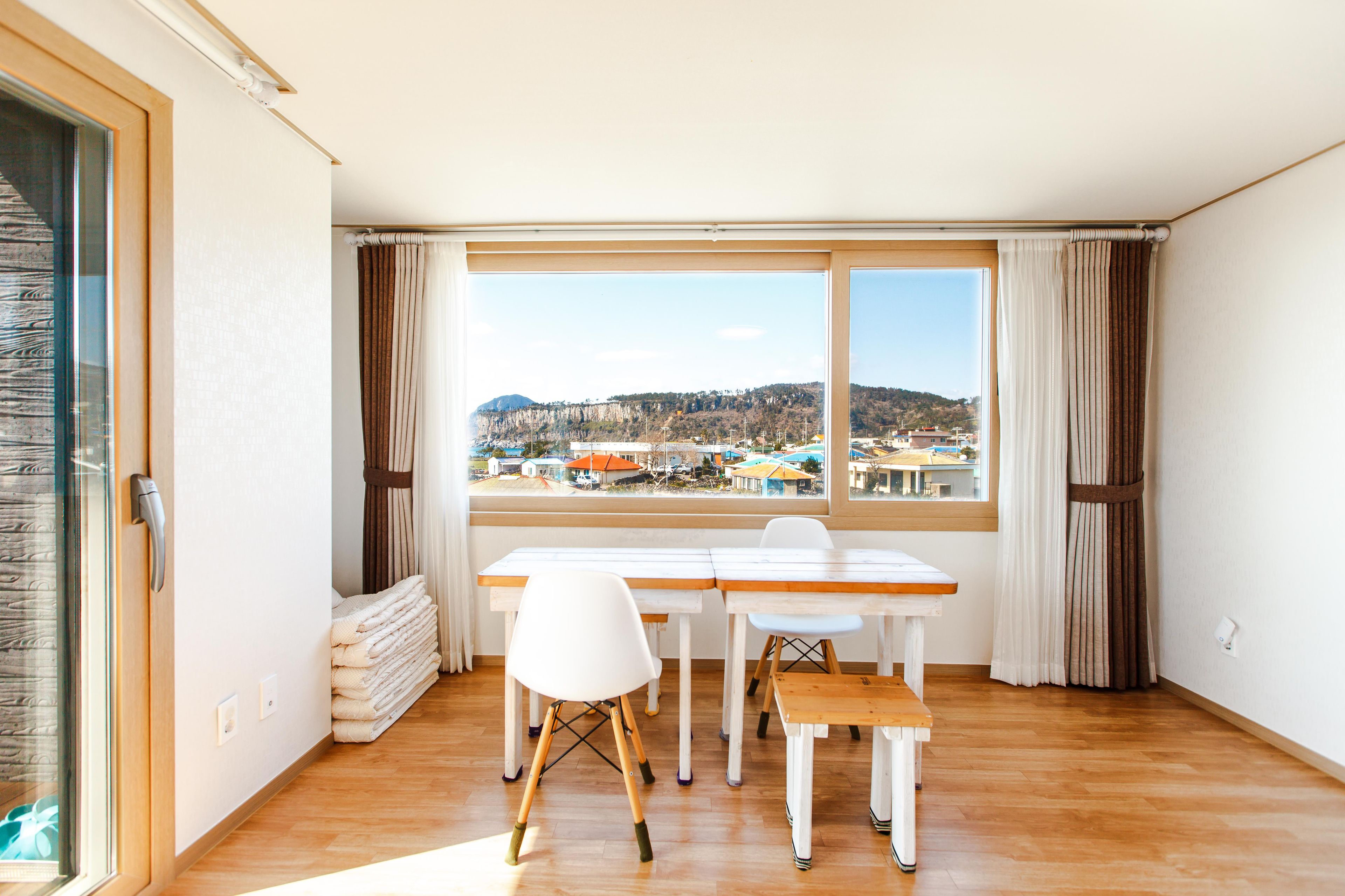 A room with a large window, wooden floor, white chairs, a wooden table, and a stack of towels.