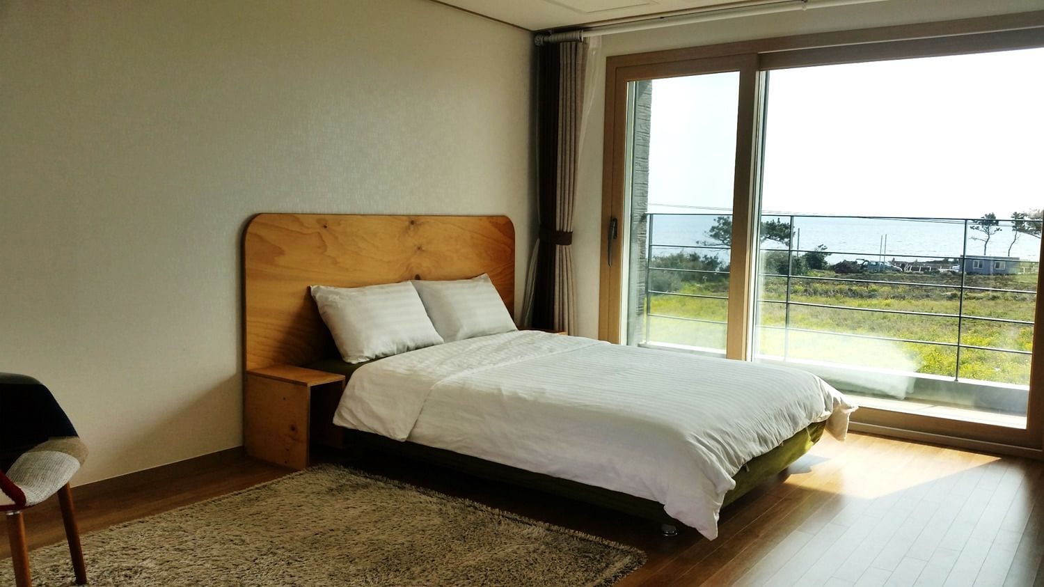 Une chambre à coucher avec une tête de lit en bois, une grande fenêtre donnant sur un paysage verdoyant et un bâtiment, et un lit recouvert de draps blancs.