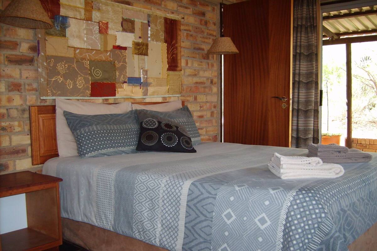 Une chambre avec un mur en briques, un lit avec une literie à motifs, une table de chevet en bois et une fenêtre avec des rideaux.