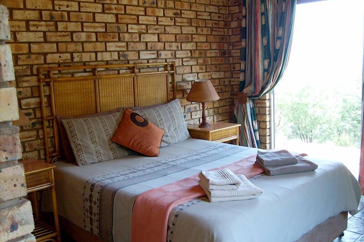 Un lit avec une tête de lit en bois, une couvre-lit à motifs et un oreiller. Il y a une table de chevet avec une lampe et une serviette pliée. La pièce a un mur en briques et une fenêtre avec des rideaux.